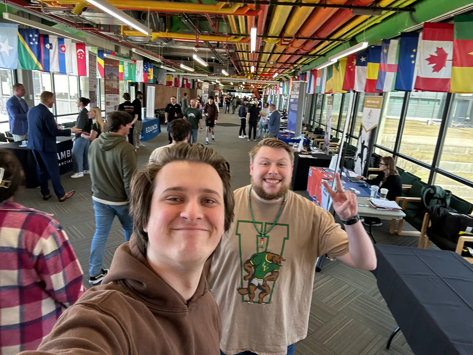 Two students smiling and posing for a selfie at a university career fair, surrounded by tables, recruiters, and international flags overhead.